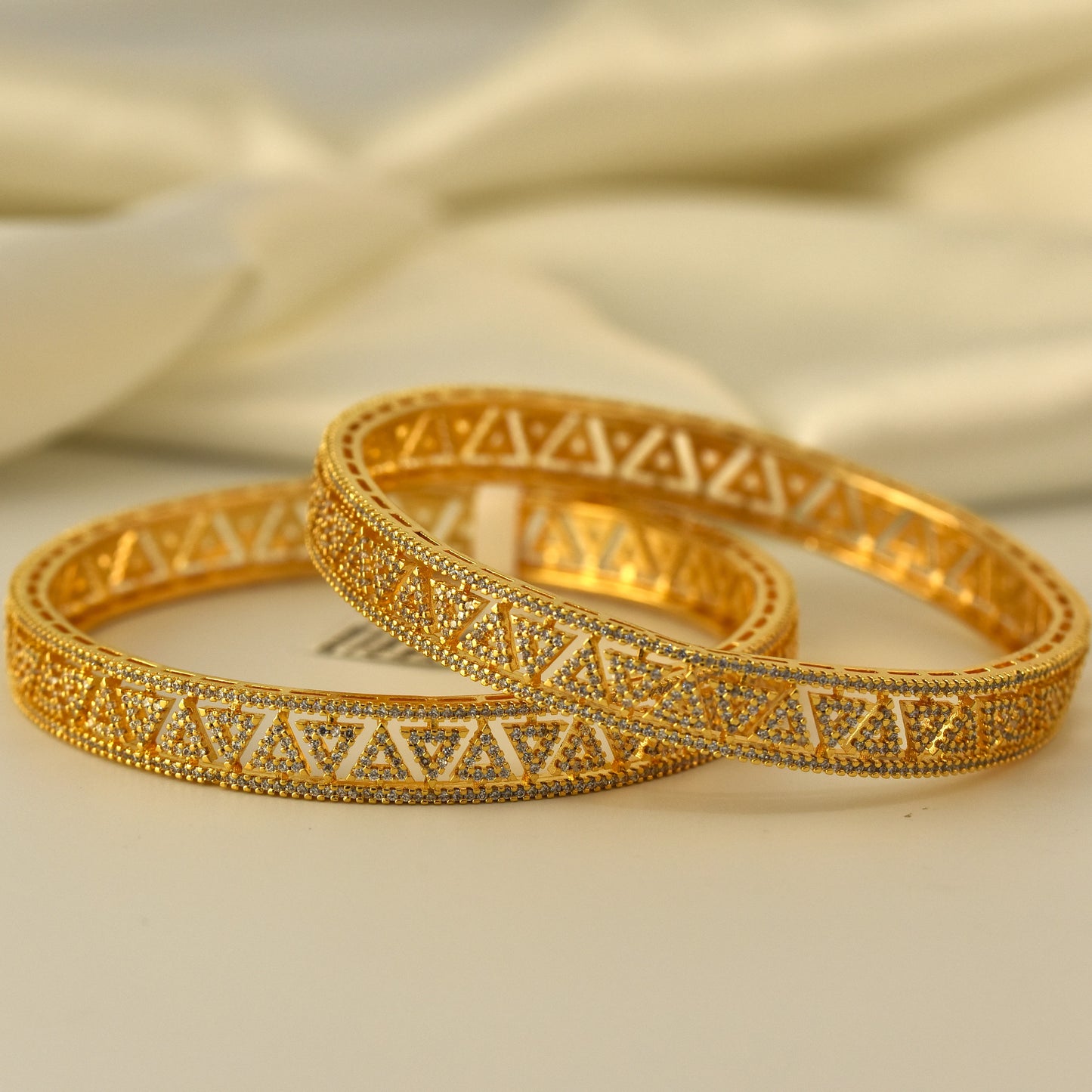 Two gold polish AD bangles with white stone intricate designs on a beige fabric background