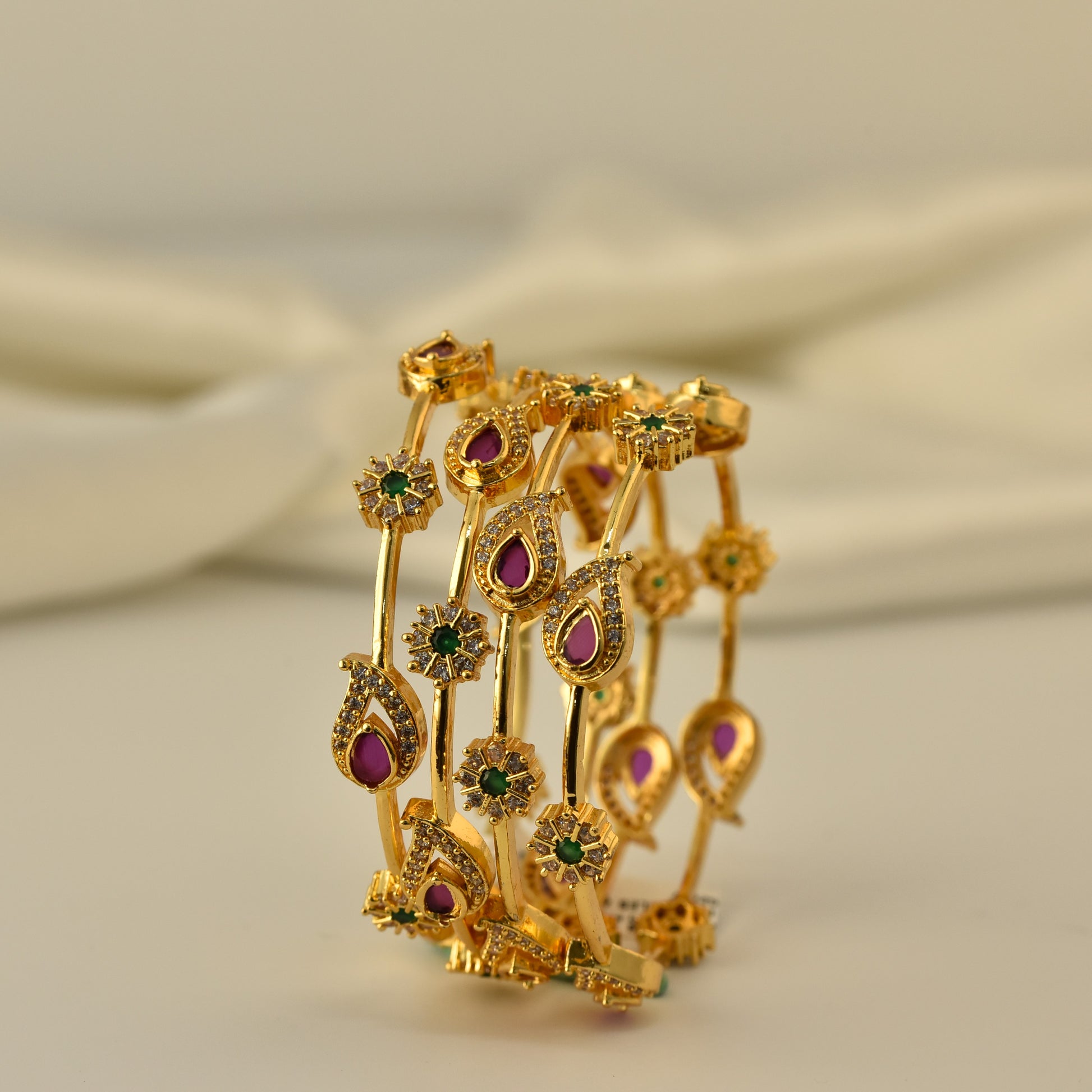 Gold polish 4pc bangle with red green and white stones in a mango shape motif on a beige background